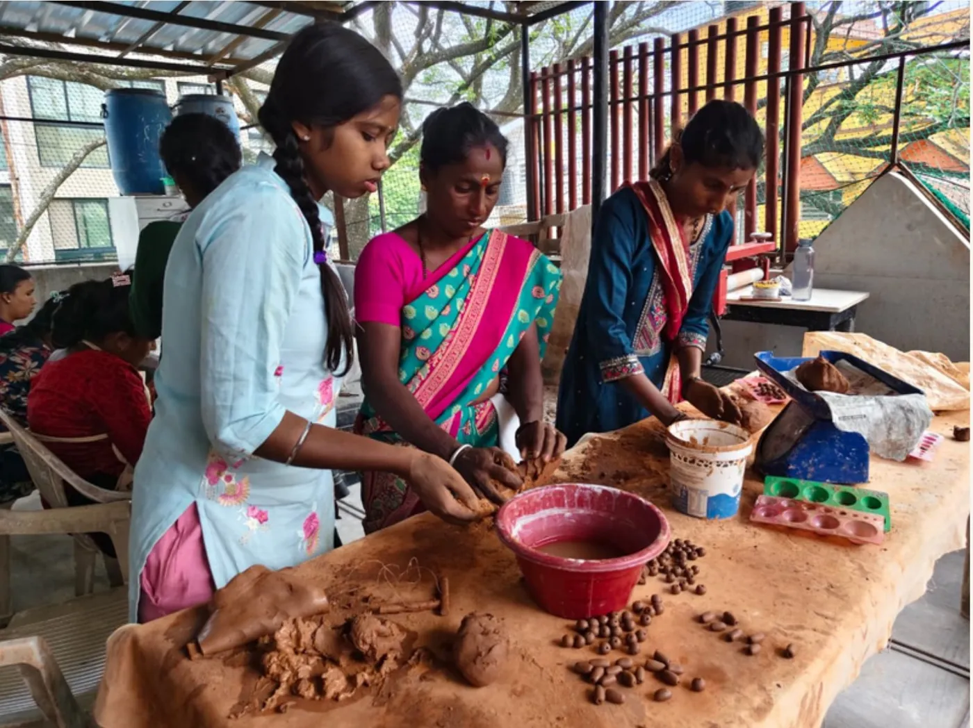 Three women shaping clay at a training table, with a bowl of slip and rows of clay beads in front of them