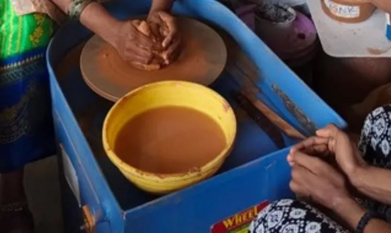 Hands centring clay on a pottery wheel beside a bowl of water, as a learner watches